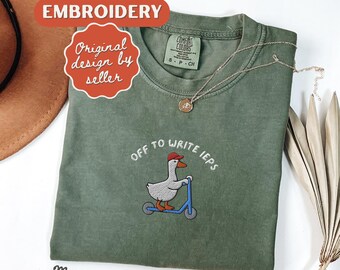 May include: Moss green t-shirt with embroidered design. The design features a goose wearing a red cap riding a blue scooter, with the text "off to write IEPs". A gold necklace is draped on the shirt.