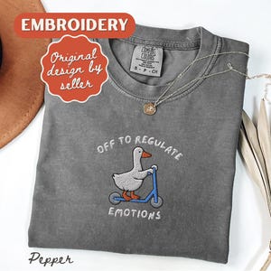May include: A gray Comfort Colors t-shirt with an embroidered design of a duck on a scooter, with the text "OFF TO REGULATE EMOTIONS". The shirt also has a small tag and a necklace.