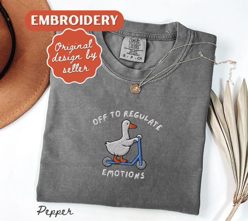 May include: A gray Comfort Colors t-shirt with an embroidered design of a duck on a scooter, with the text "OFF TO REGULATE EMOTIONS". The shirt also has a small tag and a necklace.