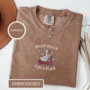 May include: Espresso-colored t-shirt with an embroidered design featuring a duck riding a scooter, the words "Busy Dilly Dallying," and small flowers. The shirt has a crew neck and a Comfort Colors tag.