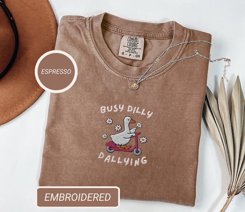 May include: Espresso-colored t-shirt with an embroidered design featuring a duck riding a scooter, the words "Busy Dilly Dallying," and small flowers. The shirt has a crew neck and a Comfort Colors tag.