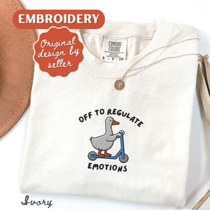 May include: Ivory-colored t-shirt with an embroidered design of a gray goose riding a blue scooter. The text reads "OFF TO REGULATE EMOTIONS." The shirt also has a gold necklace and the words "Embroidery" and "Original design by seller."