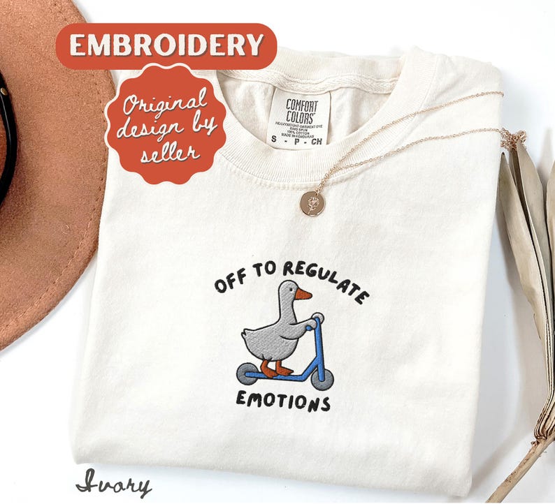 May include: Ivory-colored t-shirt with an embroidered design of a gray goose riding a blue scooter. The text reads "OFF TO REGULATE EMOTIONS." The shirt also has a gold necklace and the words "Embroidery" and "Original design by seller."