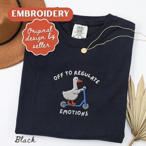 May include: Black crew neck sweatshirt with an embroidered design of a goose on a scooter, with the text "OFF TO REGULATE EMOTIONS". A gold necklace is draped on the sweatshirt. The word "Embroidery" is in a red banner.
