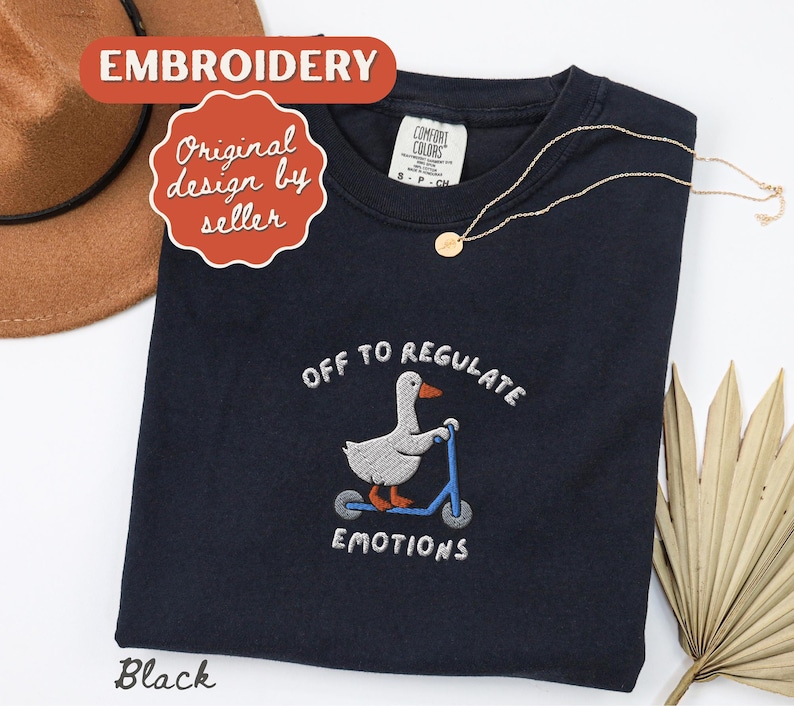 May include: Black crew neck sweatshirt with an embroidered design of a goose on a scooter, with the text "OFF TO REGULATE EMOTIONS". A gold necklace is draped on the sweatshirt. The word "Embroidery" is in a red banner.