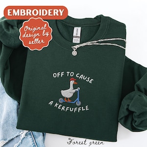 Peut inclure: Sweat-shirt vert forêt avec une broderie d'une oie sur un scooter et le texte "OFF TO CAUSE A KERFUFFLE". Un collier argenté est également visible. Le mot "Broderie" est en haut.