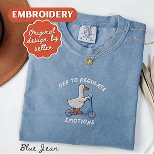 May include: A blue jean colored t-shirt with an embroidered design of a duck riding a scooter, with the text "OFF TO REGULATE EMOTIONS". The shirt also has the text "EMBROIDERY" and "Original design by seller". A gold necklace is draped across the shirt.