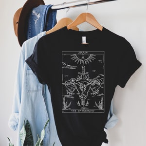Op de afbeelding: Zwart T-shirt met een witte afbeelding van een baarmoeder met een zwaard erdoorheen. De afbeelding is omringd door bloemen en planten. De tekst "Smash the Patriarchy" staat onder de afbeelding.