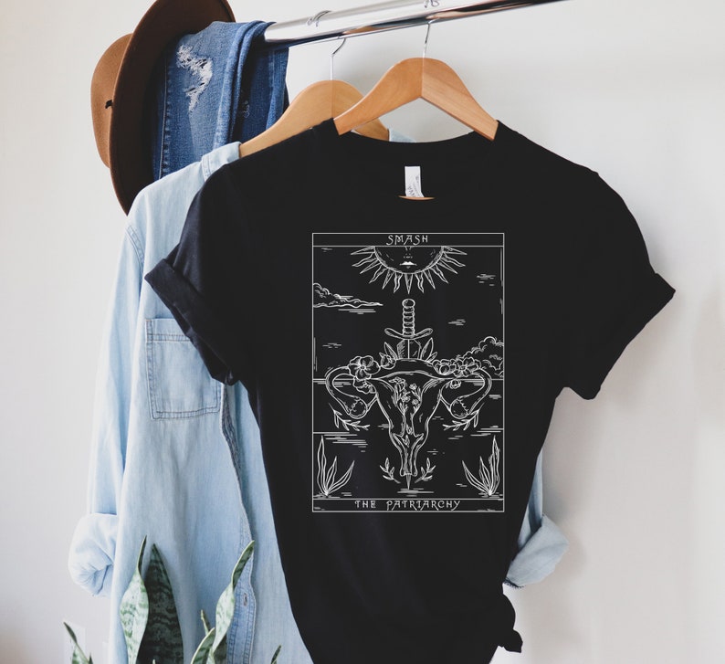 Op de afbeelding: Zwart T-shirt met een witte afbeelding van een baarmoeder met een zwaard erdoorheen. De afbeelding is omringd door bloemen en planten. De tekst "Smash the Patriarchy" staat onder de afbeelding.