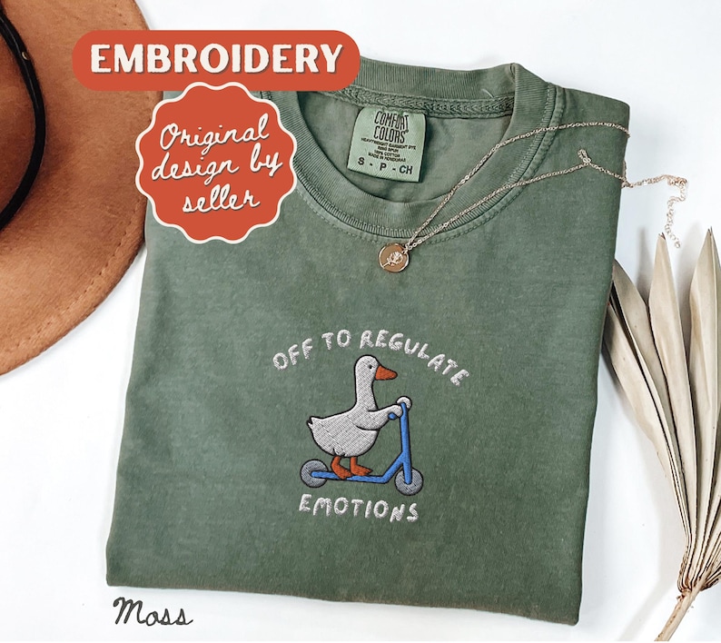 May include: A moss green t-shirt with the embroidered phrase "OFF TO REGULATE EMOTIONS" and a cartoon duck riding a blue scooter. The shirt has a crew neck and short sleeves. A gold necklace with a pendant is draped on the shirt.