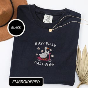May include: Black embroidered sweatshirt with the text "Busy Dilly Dallying" and an embroidered graphic of a goose riding a scooter. A gold necklace and a brown hat are also in the image. The sweatshirt is labeled "Comfort Colors".