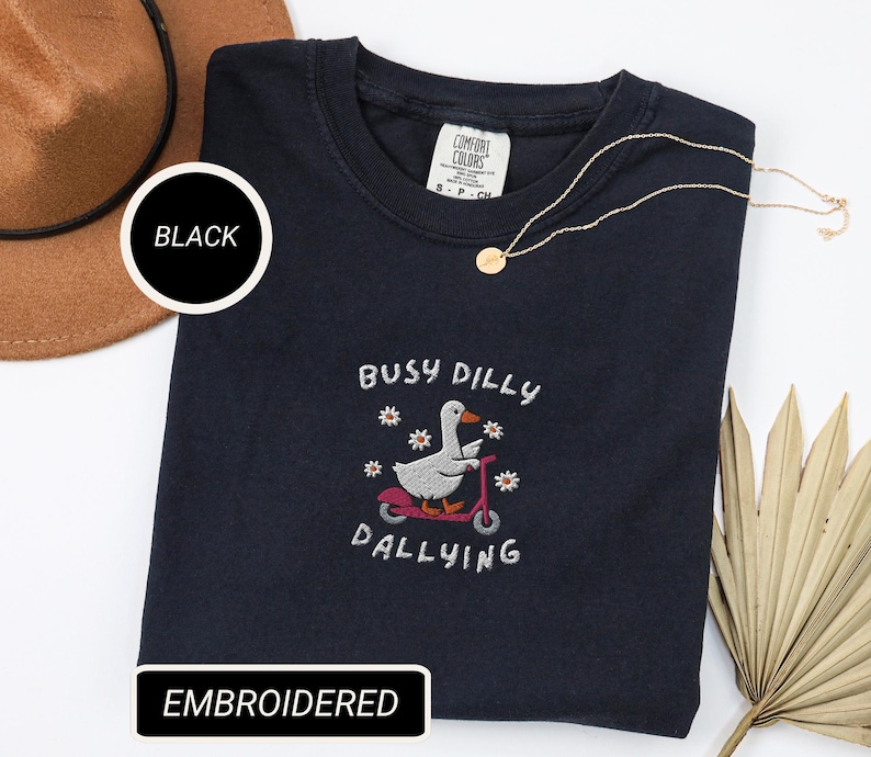 May include: Black embroidered sweatshirt with the text "Busy Dilly Dallying" and an embroidered graphic of a goose riding a scooter. A gold necklace and a brown hat are also in the image. The sweatshirt is labeled "Comfort Colors".