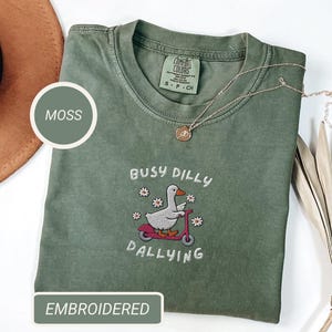May include: Moss green t-shirt with an embroidered design of a duck riding a scooter, surrounded by flowers, and the words "Busy Dilly Dallying". The shirt has a crew neck and short sleeves. A gold necklace is draped across the shirt.
