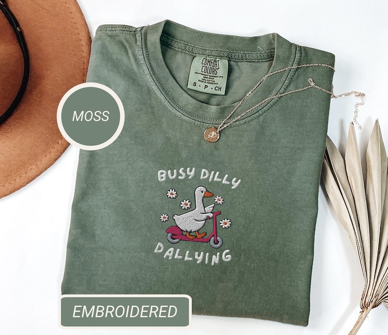 May include: Moss green t-shirt with an embroidered design of a duck riding a scooter, surrounded by flowers, and the words "Busy Dilly Dallying". The shirt has a crew neck and short sleeves. A gold necklace is draped across the shirt.