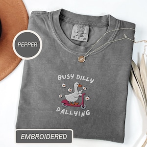 May include: A gray Comfort Colors t-shirt with an embroidered design. The design features a white goose riding a pink scooter, surrounded by flowers, and the text "BUSY DILLY DALLYING". A gold necklace and a brown hat are also visible.