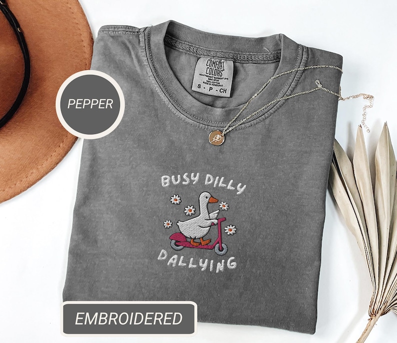 May include: A gray Comfort Colors t-shirt with an embroidered design. The design features a white goose riding a pink scooter, surrounded by flowers, and the text "BUSY DILLY DALLYING". A gold necklace and a brown hat are also visible.