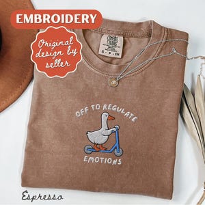 May include: Espresso-colored t-shirt with embroidered design. The design features a duck riding a blue scooter with the text "OFF TO REGULATE EMOTIONS". A silver necklace with a pendant is also visible.