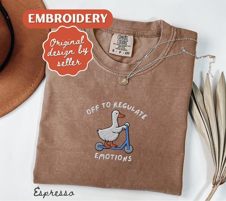 May include: Espresso-colored t-shirt with embroidered design. The design features a duck riding a blue scooter with the text "OFF TO REGULATE EMOTIONS". A silver necklace with a pendant is also visible.