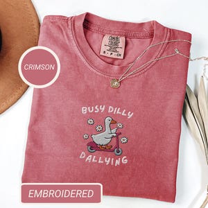 May include: Crimson-colored t-shirt with the embroidered phrase "Busy Dilly Dallying" and a cartoon duck riding a scooter. The shirt has a crew neck and short sleeves. A gold necklace is draped across the shirt.