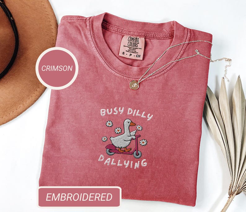 May include: Crimson-colored t-shirt with the embroidered phrase "Busy Dilly Dallying" and a cartoon duck riding a scooter. The shirt has a crew neck and short sleeves. A gold necklace is draped across the shirt.