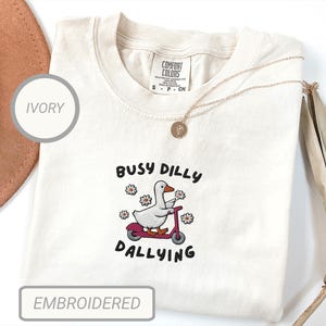 May include: Ivory-colored Comfort Colors t-shirt with an embroidered design. The design features a white goose riding a red scooter, surrounded by flowers, with the text "Busy Dilly Dallying". The shirt has a crew neck and short sleeves.