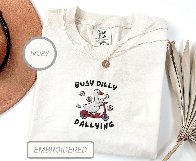 May include: Ivory-colored Comfort Colors t-shirt with an embroidered design. The design features a white goose riding a red scooter, surrounded by flowers, with the text "Busy Dilly Dallying". The shirt has a crew neck and short sleeves.