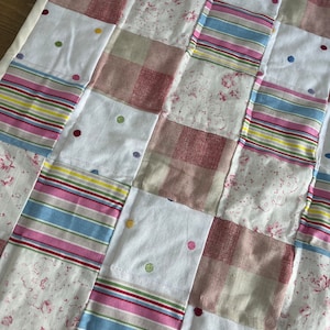 May include: A patchwork quilt featuring a mix of fabrics, including stripes, floral prints, and gingham. The quilt is made in a range of colours, including pink, blue, white, and beige. The quilt is decorated with small, colourful dots.