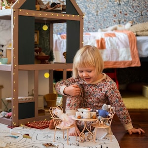 Op de afbeelding: Een jong meisje in een pyjama met bloemenprint zit op een wit tapijt met een speelgoed theeservies. Het theeservies bevat een theepot, twee kopjes, een bord en een suikerpot. Het tapijt heeft een zwart-wit ontwerp van een stad met huizen, wegen en bomen.