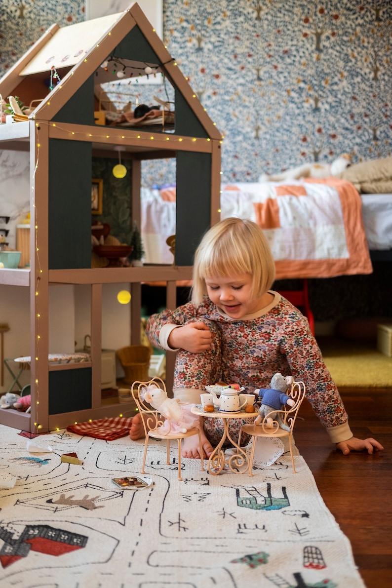 Op de afbeelding: Een jong meisje in een pyjama met bloemenprint zit op een wit tapijt met een speelgoed theeservies. Het theeservies bevat een theepot, twee kopjes, een bord en een suikerpot. Het tapijt heeft een zwart-wit ontwerp van een stad met huizen, wegen en bomen.