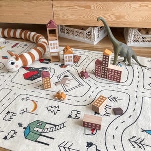 May include: A cream-colored play mat featuring black line drawings of roads, trees, and buildings, accompanied by a plush brown and white striped caterpillar, several wooden building blocks, small wooden toy cars, and a green dinosaur figure.