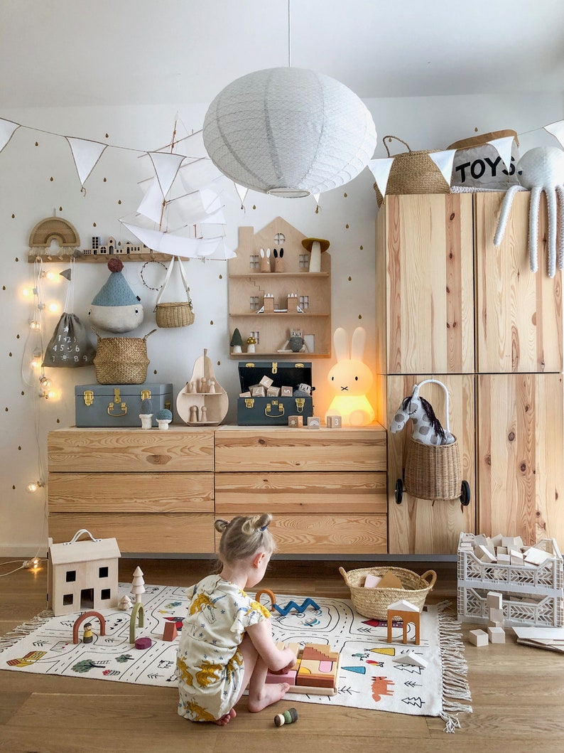 Op de afbeelding: Een kind speelt met houten blokken op een speelkleed met een stadsontwerp. Het kleed is wit met zwarte contouren en toont een weg, huizen, bomen en andere stedelijke elementen. Het kind zit op het kleed en draagt een geel-witte jurk. De kamer is versierd met een witte papieren lantaarn, een houten plank met speelgoed en een houten kast met een bordje waarop "TOYS" staat.