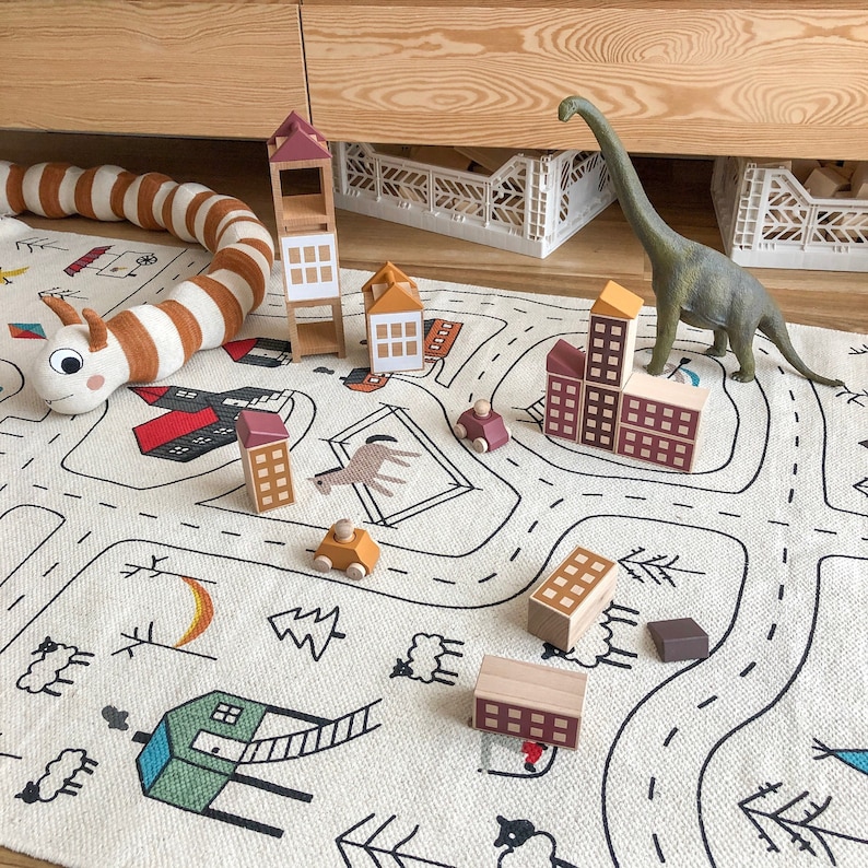 May include: A white play mat with black lines forming a road and a town.  There are wooden toy houses, cars, and a dinosaur on the mat.