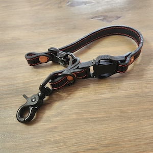 May include: Black leather camera strap with red stitching and copper accents. The strap has a black plastic buckle and a black metal clip.