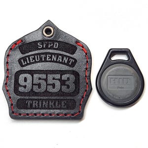 Peut inclure: Porte-clés en cuir noir avec des coutures rouges en forme de casque de pompier. Le porte-clés porte la gravure "SFPO LIEUTENANT 9553 TRINKLE". Un petit porte-clés noir avec une carte HID Prox grise est également présenté.