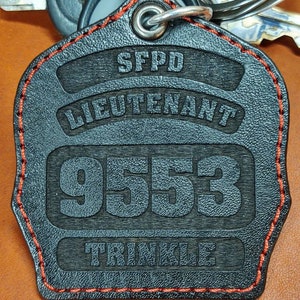 May include: Black leather keychain with red stitching. It has the text "SFPD Lieutenant 9553 Trinkle" engraved on it.