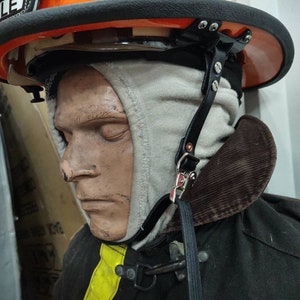 May include: An orange firefighter helmet with a black brim and a leather strap. The helmet is worn by a mannequin wearing a tan hood and a black coat with a yellow stripe.
