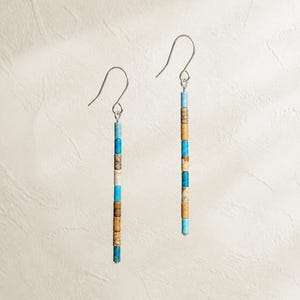 May include: A pair of dangle earrings featuring long, thin vertical columns of alternating cylindrical beads. The beads are colored in shades of bright turquoise, tan, and cream with speckled textures, suspended from simple silver-toned metal hooks.