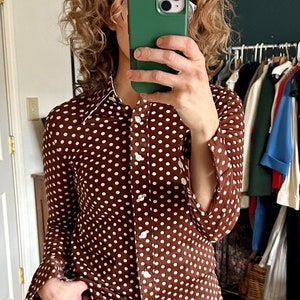 Op de afbeelding: Een vrouw draagt een bruin en wit geblokt shirt met een witte kraag en blauwe jeans met gescheurde knieën.