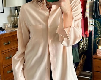 May include: A pale pink button-down shirt with long sleeves and a chest pocket. The shirt is made of a silky material and has a relaxed fit.