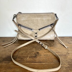 May include: A beige leather crossbody bag featuring a top zipper closure, two diagonal front zippered pockets, and two decorative metal studs. The bag includes a long, adjustable shoulder strap with metal hardware and long leather zipper pulls.