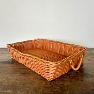 May include: Rectangular woven wicker basket in a warm orange hue. The basket features a woven design with a rectangular shape and curved handles on each end. The basket is suitable for storage or display.