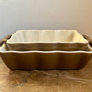 Puede incluir: Un juego de dos fuentes para hornear de cerámica marrón con bordes festoneados y asas doradas. La fuente más pequeña está anidada dentro de la fuente más grande.