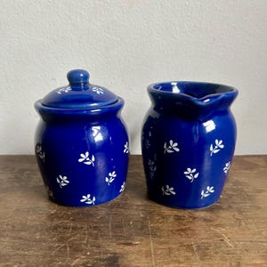 May include: A blue and white ceramic creamer and sugar bowl set with a floral pattern. The creamer has a handle and a spout. The sugar bowl has a lid.