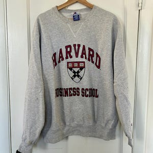 Pode incluir: Um moletom cinza claro da Harvard Business School com gola redonda e mangas compridas. O moletom apresenta a palavra "HARVARD" em letras arqueadas bord&ocirc; acima de um bras&atilde;o e "BUSINESS SCHOOL" abaixo.