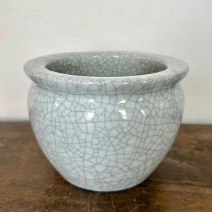 May include: A round, light blue ceramic planter with a crackle glaze finish. The planter has a wide, rounded rim and a bulbous body. The surface is covered in a network of fine, dark gray lines, creating a unique texture.