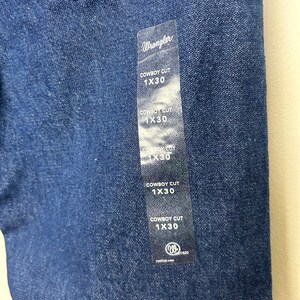 Puede incluir: Primer plano de unos vaqueros de mezclilla azul oscuro con una etiqueta Wrangler. La etiqueta dice "COWBOY CUT 1X30" varias veces. Los vaqueros son de estilo cl&aacute;sico, con costuras visibles y una tela duradera.