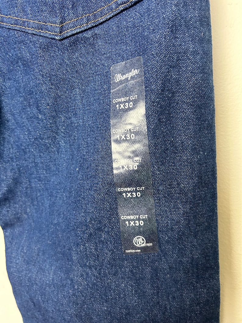 Puede incluir: Primer plano de unos vaqueros de mezclilla azul oscuro con una etiqueta Wrangler. La etiqueta dice "COWBOY CUT 1X30" varias veces. Los vaqueros son de estilo cl&aacute;sico, con costuras visibles y una tela duradera.