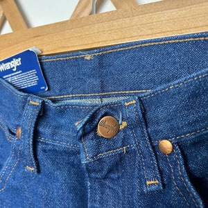Puede incluir: Primer plano de unos vaqueros Wrangler de mezclilla azul. Los vaqueros tienen un cierre de bot&oacute;n con el logotipo de Wrangler, remaches de cobre y una etiqueta azul con texto blanco. Los vaqueros est&aacute;n colgados de una percha de madera.