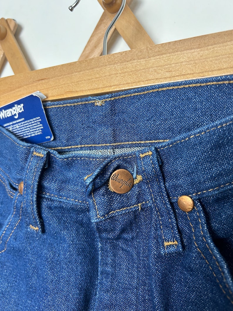 Puede incluir: Primer plano de unos vaqueros Wrangler de mezclilla azul. Los vaqueros tienen un cierre de bot&oacute;n con el logotipo de Wrangler, remaches de cobre y una etiqueta azul con texto blanco. Los vaqueros est&aacute;n colgados de una percha de madera.
