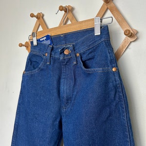Puede incluir: Vaqueros Wrangler azul oscuro colgados en un perchero de madera. Los vaqueros tienen un dise&ntilde;o cl&aacute;sico de cinco bolsillos con remaches color cobre y cierre de bot&oacute;n. Una etiqueta azul con el logotipo de Wrangler est&aacute; adjunta.